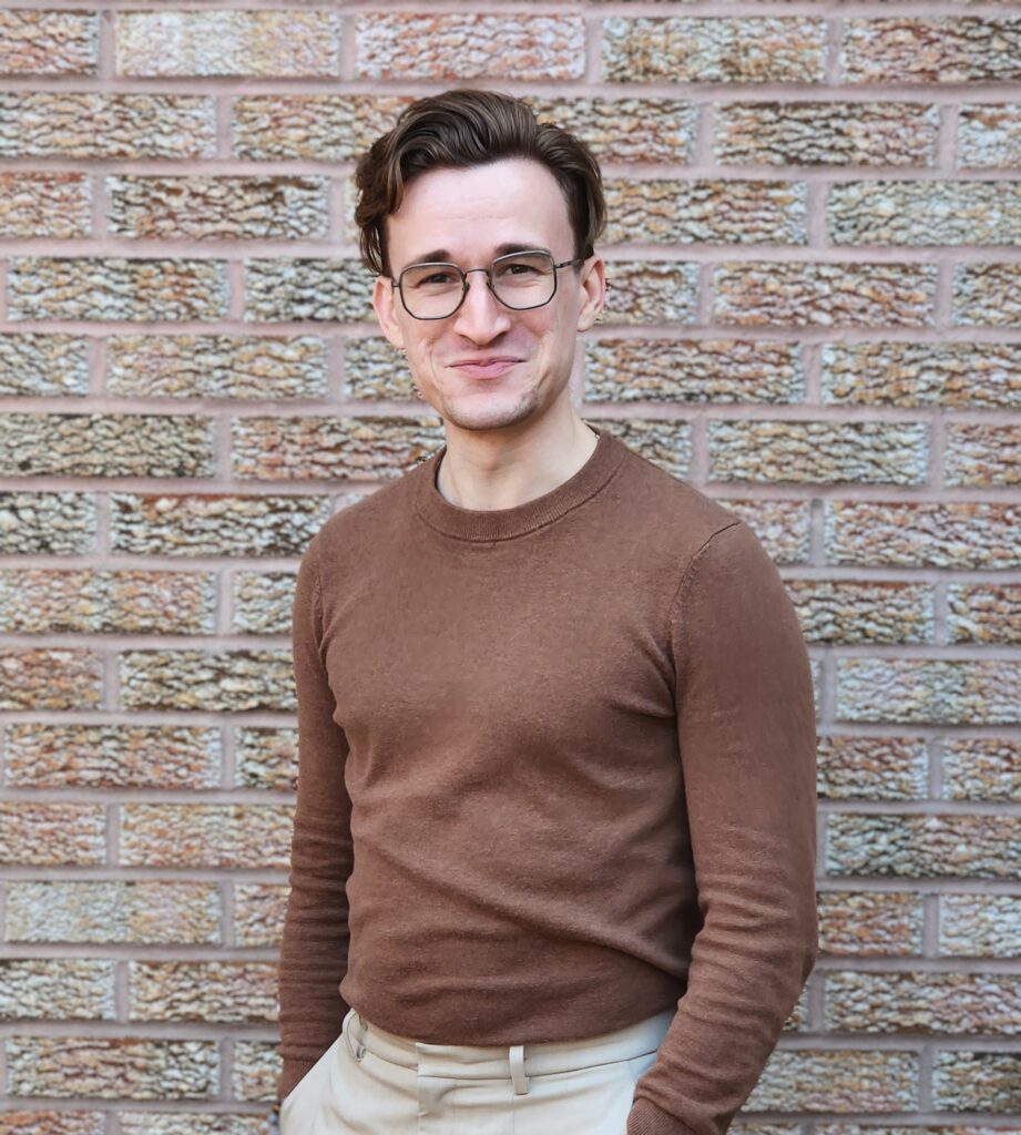 Phill, wearing glasses, in a brown sweater, against a brick wall, smiling at the camera.